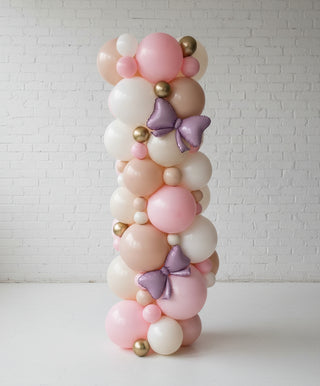 A decorative balloon column with a organic design, featuring a variety of colors including pink, white, and beige, adorned with purple bows and gold foil accents.