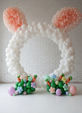 A decorative Easter-themed hoop with balloon ears and a colorful basket of eggs and flowers in front of it.