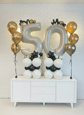 Balloon arrangement with '50' for a 50th birthday celebration on a white cabinet against a light brick wall.