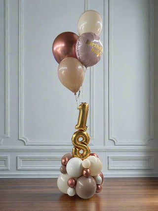 A tabletop birthday decoration featuring a large number 18 with attached helium balloons in pink, gold, and white colors, on a white background.