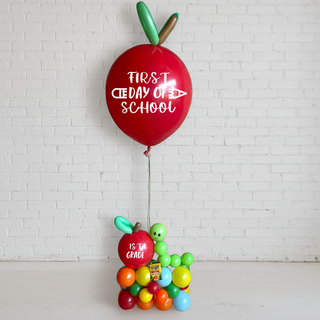 A large red balloon designed to look like an apple with 'FIRST DAY OF SCHOOL' written in white, attached to a small balloon base decorated with colorful balloons.