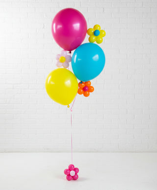 Colorful balloons with flower-shaped ribbons against a white brick wall.