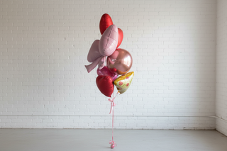 A bouquet of foil balloons shaped like pizza and pizza slices, in various shades of pink, red, and yellow, tied together with pink ribbon against a white background.