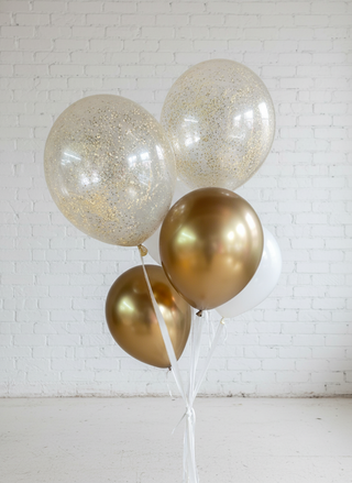 A bouquet of balloons including clear and gold colored balloons with a sparkling glittery finish, tied together with white string, standing in a room with a light background.