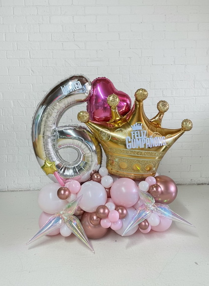 Decorative balloons with numbers, crowns, and hats on a wooden floor against a brick wall.