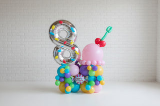 A balloon pedestal decoration featuring a large number 8, with colorful balloons in a variety of shapes and a 'Happy Birthday' message, designed to resemble a dessert theme.