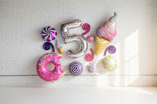 A variety of foil balloons in shapes of desserts and sweets, including a number 5, a donut, and an ice cream cone, displayed against a beige background.