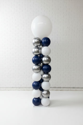 A tall balloon column decoration made of alternating white, navy blue, and metallic silver balloons, standing in a balloon shop with wood floors and other balloons around it.