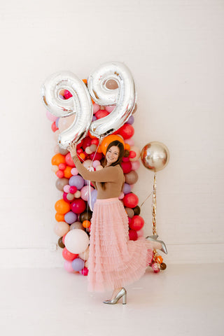 Founder of 99 Haus Balloons and Decor holding two silver metallic number balloons "99" as she walks in front of a colorful balloon column.