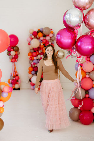 Founder of 99 Haus Balloons and Decor holding two silver metallic number balloons "99" as she walks in front of a colorful balloon column.