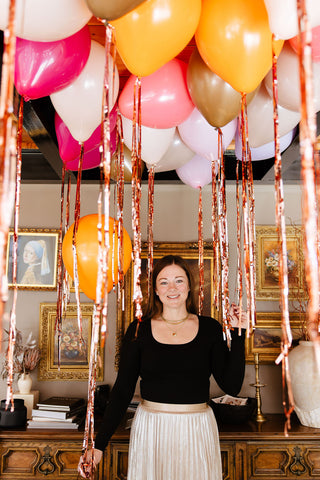 Founder of 99 Haus Balloons and Decor holding metallic balloon tassels in front of an elegant gallery wall filled with gold frames