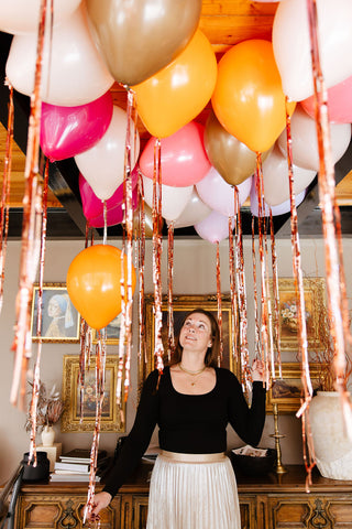 Founder of 99 Haus Balloons and Decor holding metallic balloon tassels in front of a gallery wall of golden frames