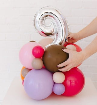 Colorful balloon arrangement with a hand adjusting a silver number balloon on a light background