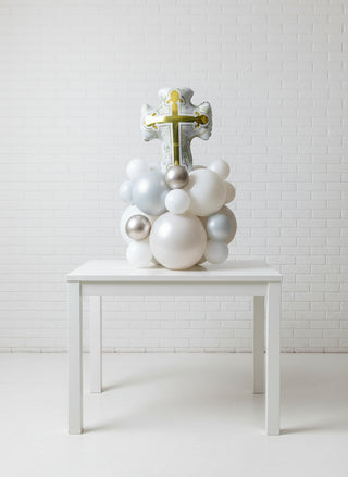 A decorative tabletop featuring a large gold cross with intricate designs, surrounded by a cluster of white and silver balloons.