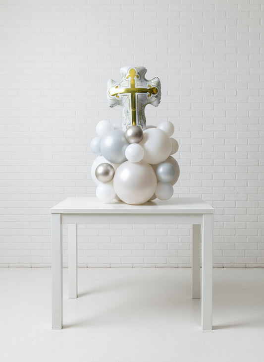A decorative tabletop featuring a large gold cross with intricate designs, surrounded by a cluster of white and silver balloons.