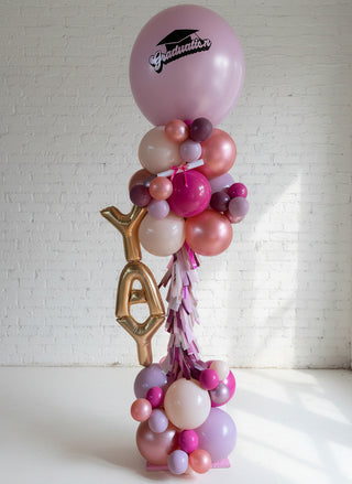 A balloon tower featuring a variety of balloons in pink, gold, and white colors, with a 'Graduation' sign at the top.