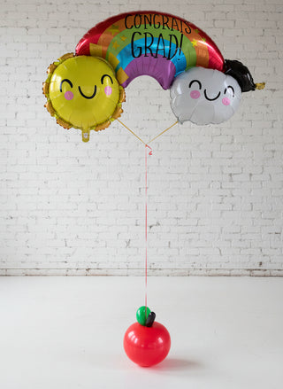A bouquet of graduation-themed balloons, featuring a large rainbow-shaped balloon with 'CONGRATS GRAD!' text, along with two character balloons and a small red balloon shaped like an apple.