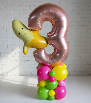A balloon pedestal with a number shape, decorated with fruit characters and filled with multicolored balloons.