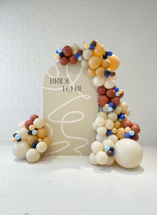 A custom event backdrop featuring the phrase 'BRIDE TO BE' surrounded by an array of multicolored balloons in shades of gold, cream, and blue, creating a festive and celebratory atmosphere.