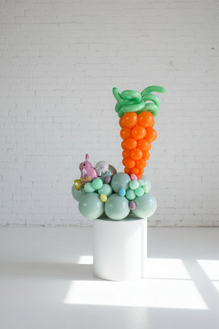 A balloon centerpiece designed to look like an Easter carrot with a top resembling a carrot, surrounded by eggs and a small plush toy rabbit.