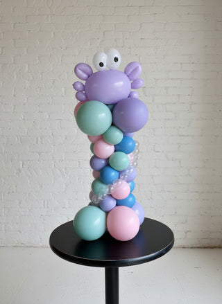 A balloon centerpiece designed to look like a deep sea creature, with a crab motif, in shades of purple, blue, and pink, displayed on a black table against a white background.
