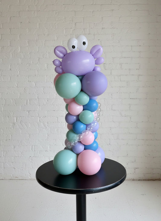 A balloon centerpiece designed to look like a deep sea creature, with a crab motif, in shades of purple, blue, and pink, displayed on a black table against a white background.