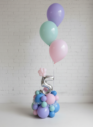 A helium centerpiece featuring balloons in shades of blue and pink, shaped into a number '5' design, arranged on a table with a plain background.