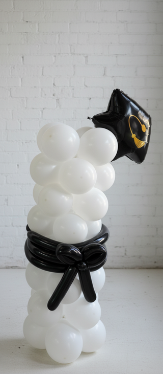 A balloon column decoration with a graduation diploma-shaped topper, predominantly white with black accents and a gold seal at the top.
