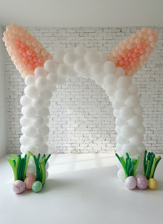 A balloon arch decoration designed to resemble Easter bunny ears, with white and pink balloons, Easter egg accents, and artificial grass at the base.