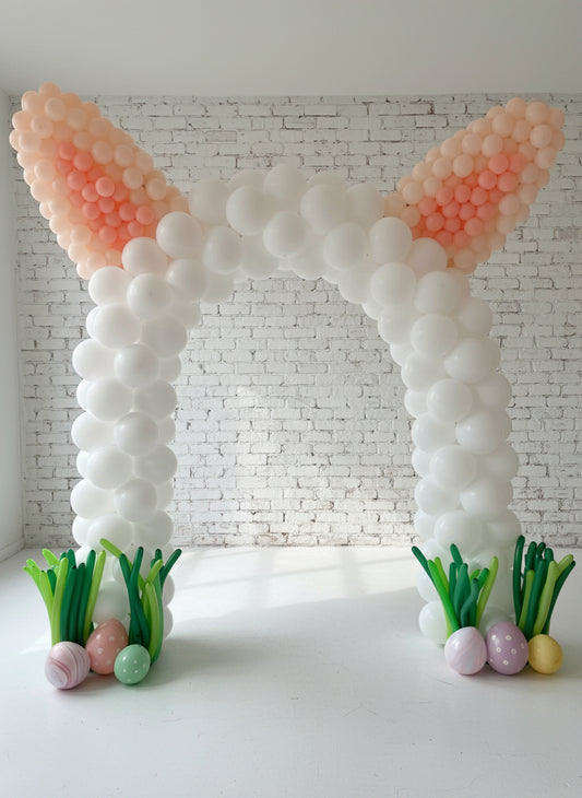 A balloon arch decoration designed to resemble Easter bunny ears, with white and pink balloons, Easter egg accents, and artificial grass at the base.