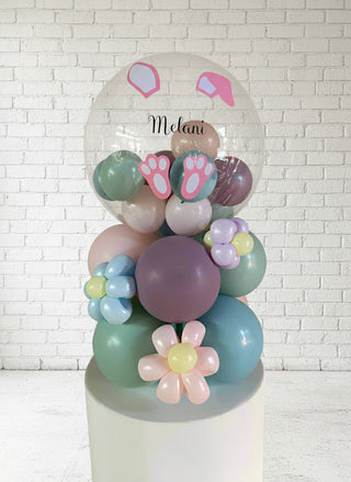 A tabletop balloon arrangement featuring a bunny design with pastel colors, including pink, purple, and green balloons, with a custom name 'Melanie' in pink lettering on the top.
