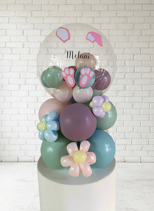 A tabletop balloon arrangement featuring a bunny design with pastel colors, including pink, purple, and green balloons, with a custom name 'Melanie' in pink lettering on the top.