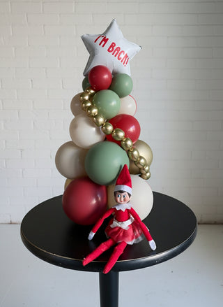A decorative Christmas tree-shaped balloon arrangement with red, green, gold, and white balloons, topped with a star, and a toy elf in a red suit sitting at the base.
