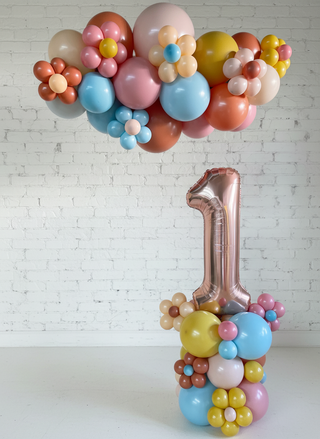 A set of balloon decorations featuring a large number 1, with a garland of multicolored balloons arranged in a floral pattern around and above it, against a white paneled wall.