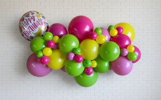An arrangement of colorful balloon garland featuring a variety of fruit colors, with larger foiled balloons in the background.