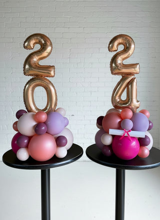Two balloon centerpiece decorations designed with large gold numbers '20' and '21' on top, surrounded by multicolored balloons in pink, purple, and red hues, displayed on black tables against a wooden background.