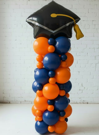 A balloon column decoration featuring a graduation cap on top, with a gradient of blue to gold balloons, representing a graduation celebration.