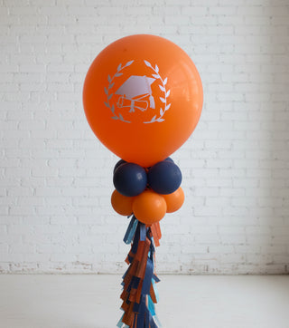 A graduation-themed centerpiece featuring an orange balloon with a graduation cap design, accompanied by smaller blue and orange balloons, and a decorative ribbon.
