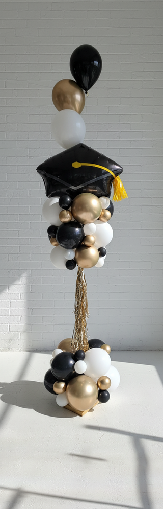 A decorative balloon tower with a graduation cap on top, featuring a mix of black, gold, white, and blue balloons, with a small balloon bouquet attached to the side.