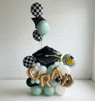 A balloon pedestal featuring a graduation theme with a large checkered graduation cap at the top, surrounded by balloons in shades of black, white, and gray, and the word 'GRAD' prominently displayed.