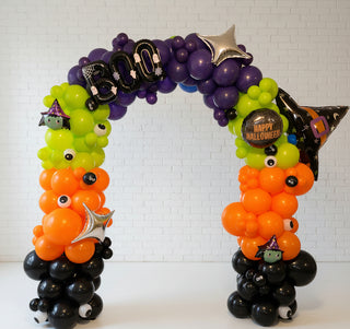 A Halloween-themed balloon arch with a variety of colors including purple, orange, and black, with decorations like a full moon, stars, and a 'Happy Halloween' sign.
