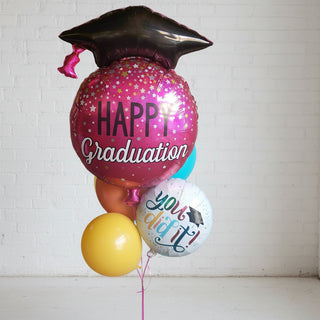 A helium balloon bouquet featuring a large pink balloon with 'HAPPY GRADUATION' text, a graduation cap, and other small balloons in a variety of colors.