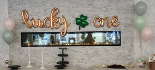 A display of St. Patrick's Day themed decorations including a 'Lucky One' sign in cursive script, with a green four-leaf clover, balloons in shades of green, gold, and white, and a decorative tray with treats.
