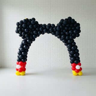A balloon arch designed to resemble Mickey Mouse ears, with a predominantly black structure, red and yellow accents at the base.