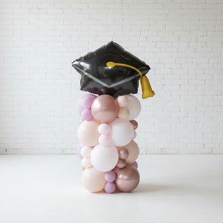 A decorative balloon column with a graduation cap on top, featuring a mix of white, silver, and gold balloons.