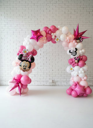 A balloon arch featuring Minnie Mouse character designs, pink and white balloons, and decorative elements like bows and polka dots, with a round table in the background.