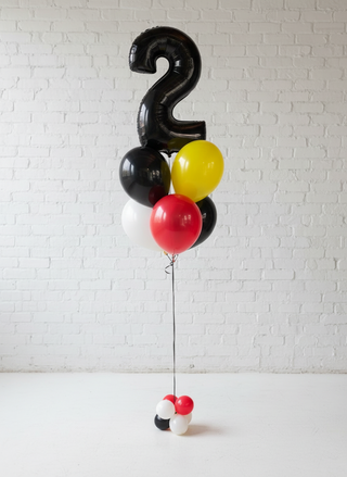 A helium balloon bouquet featuring numbers and character shapes in black, red, yellow, and white colors, designed to resemble Mickey or Minnie Mouse, placed against a white wall.