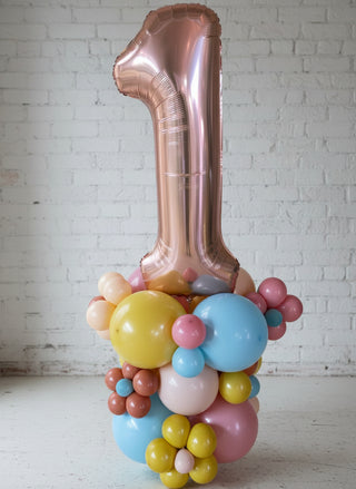 A decorative balloon column with the number 1 shape in a metallic rose gold color, surrounded and partially covered by smaller multicolored balloons in yellow, blue, pink, and other colors.