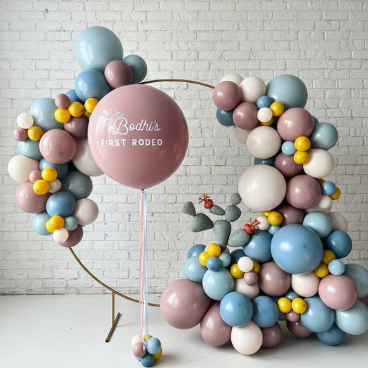 A hoop backdrop decorated with a variety of balloons in different colors, featuring a large balloon with the text 'Bobbi's First Rodeo'.
