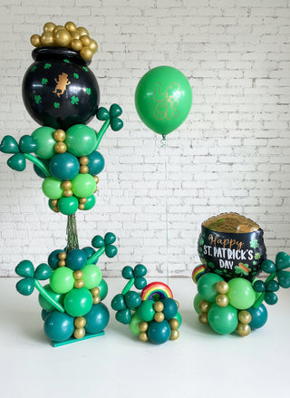 A collection of St. Patrick's Day themed balloons, featuring a large green column with gold accents, alongside a green balloon with a face, and two smaller table top decorations with clover designs.